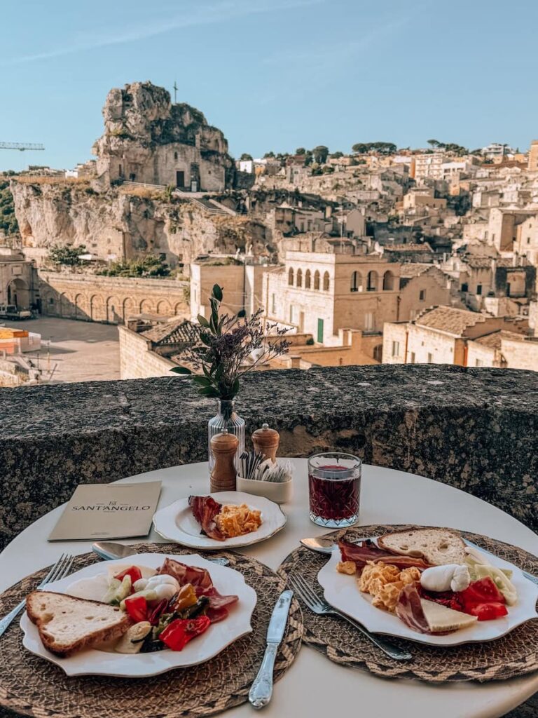 Breakfast on the terrace at Sant'Angelo Luxury Resort in Matera, Italy, with plates of eggs, cheese, cured meats, fresh vegetables, and bread overlooking the Sassi district and the rock-hewn Church of San Pietro Caveoso. 