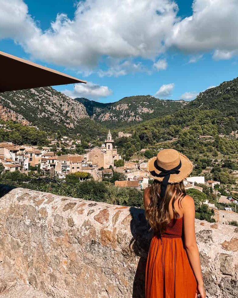 Woman in a rust-colored sundress and straw hat with a black ribbon, standing on a stone terrace overlooking the village of Valldemossa in Mallorca, Spain. The view includes terracotta rooftops, a church with a bell tower, and lush green mountains under a bright blue sky with scattered white clouds.